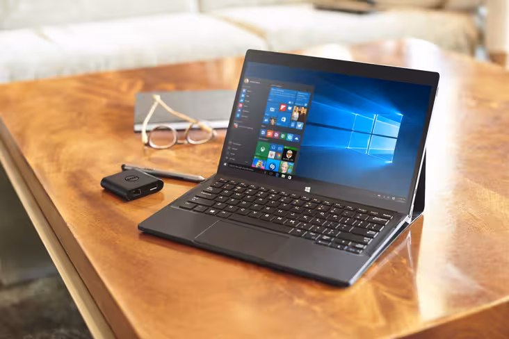 Dell XPS 12 (Model 9250) 2-in-1 notebook computer with Premier Folio, Dell USB Adapter Type-C HDMI/VGA/Ethernet/USB 3.0 (Slayer), and Active Stylus (Juniper) on a wooden coffee table in a bright lobby with a pair of glasses and notebook beside it. A tan c