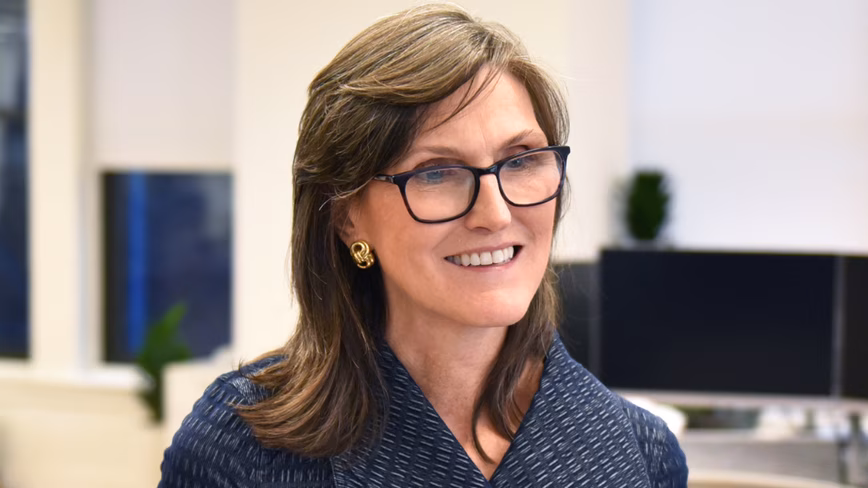 Cathie Wood, in a dark blue jacket and glasses, stands smiling in an open-plan office holding a sheet of paper.