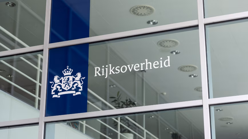 The Dutch central government's Rijksoverheid logo and coat of arms on a window of a government building, with a staircase visible inside.