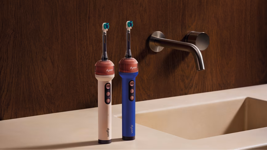 Two Dyson CameraJet electric toothbrushes, one cream and one blue, standing on a bathroom counter beside a wall-mounted tap