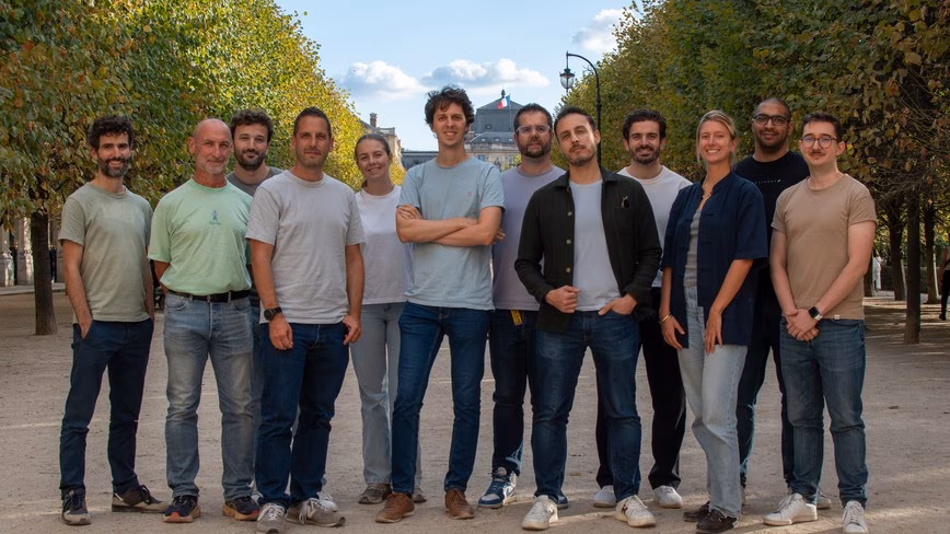 Twelve people in casual clothes stand in a row on a gravel path between clipped trees, with a building flying the French flag behind them.