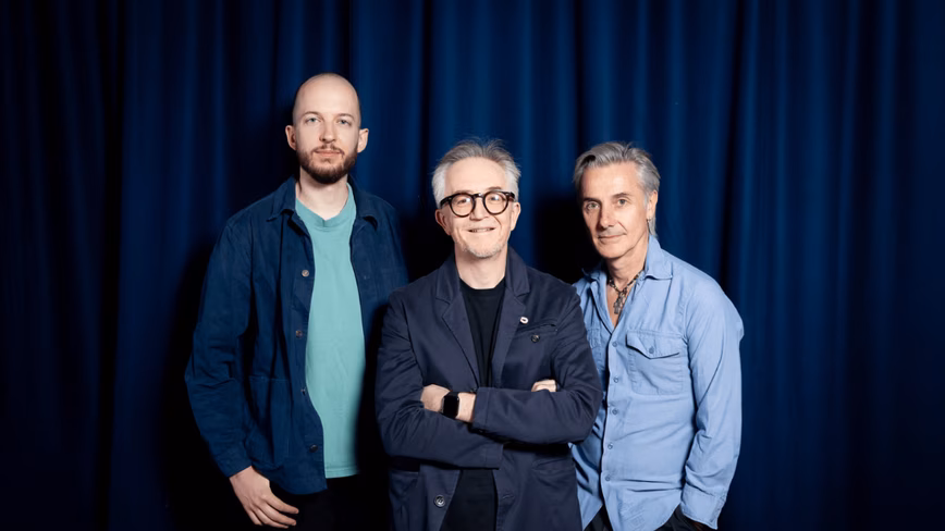 Pavle Mihajlovic, Barney Wragg and Jonathan Butterell of Unit1 Studio, standing in front of a dark blue curtain.