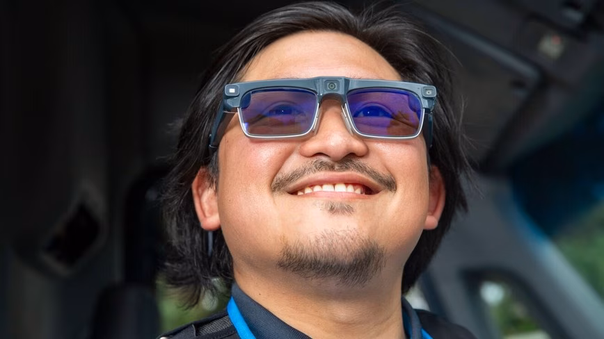 Smiling delivery driver in a blue lanyard wearing grey smart glasses with small cameras on the frame, seated in a van