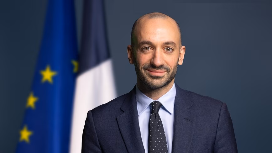 Benjamin Haddad in a navy suit and dotted tie, smiling in front of EU and French flags.