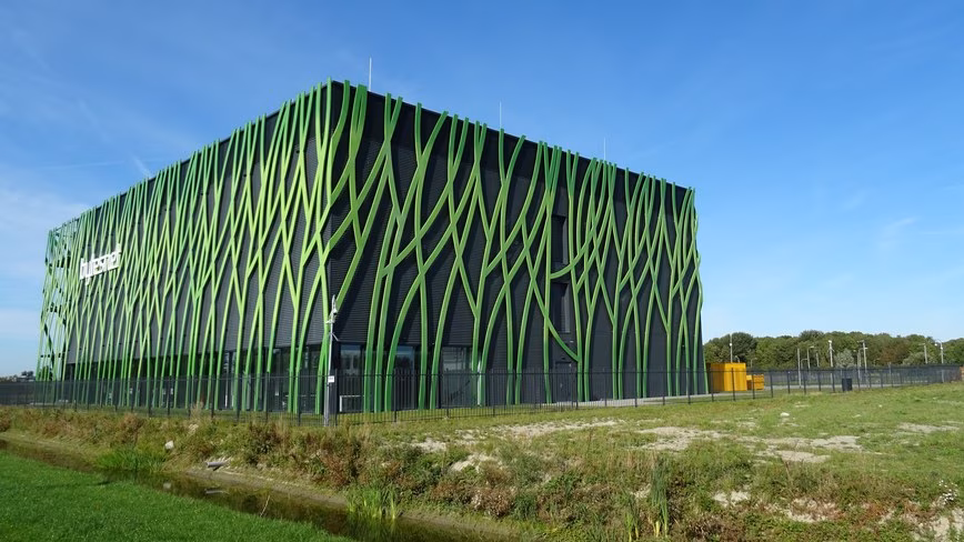 Black cube-shaped data centre wrapped in a lattice of green branching steel ribs, behind a fence and a grassy ditch.