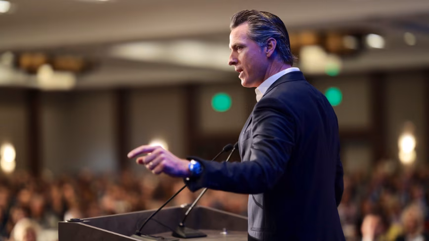 Gavin Newsom speaking at a lectern, gesturing towards an audience in a conference hall.