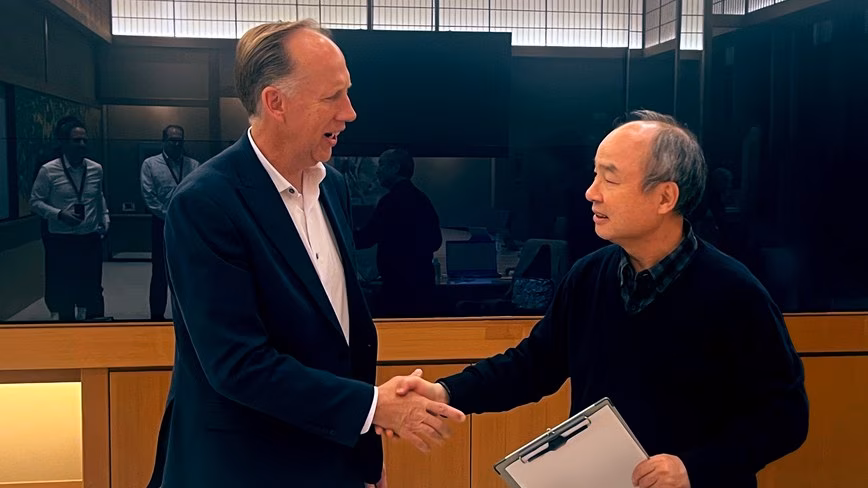 Two men shake hands in a wood-panelled meeting room, one in a navy blazer, the other in a dark jumper holding a clipboard.