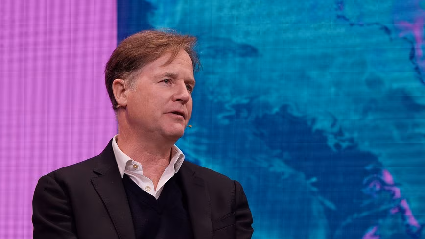 Nick Clegg, in a dark jacket and open-collared shirt, speaks on stage in front of a large pink and blue screen.