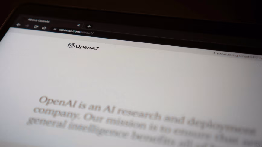 Close-up of a laptop screen showing the OpenAI logo and the openai.com about page, its mission statement blurred in the foreground