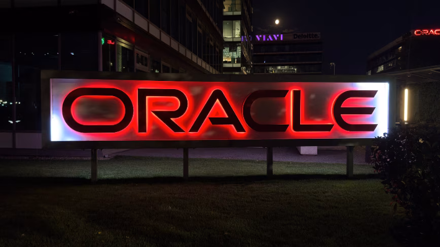 An illuminated red Oracle sign stands on a lawn at night, with lit office buildings behind it.