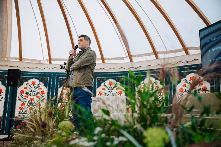 Philip Lakin of Zapier speaking into a microphone in a yurt with floral wall panels, behind tall grasses and flowers