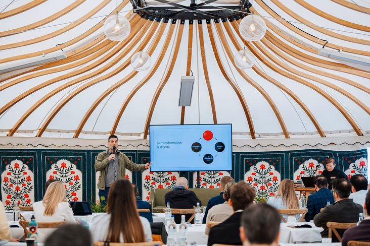 Philip Lakin of Zapier speaking into a microphone beside a screen reading AI transformation is a loop, in front of seated attendees in a tent
