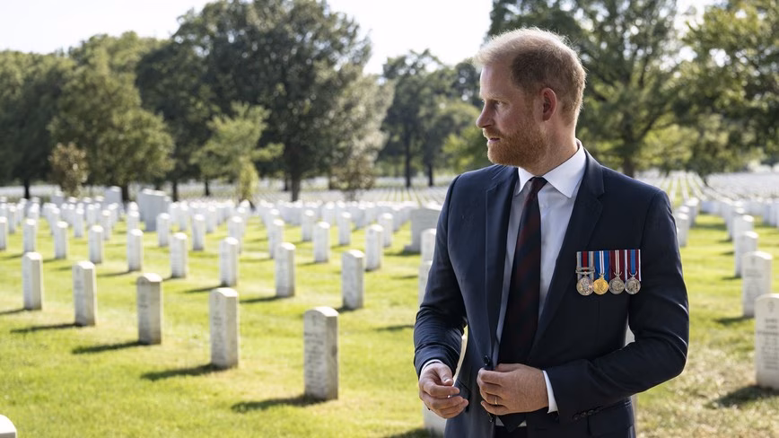 Prince Harry in a dark suit with four medals looks across rows of white headstones at Arlington National Cemetery
