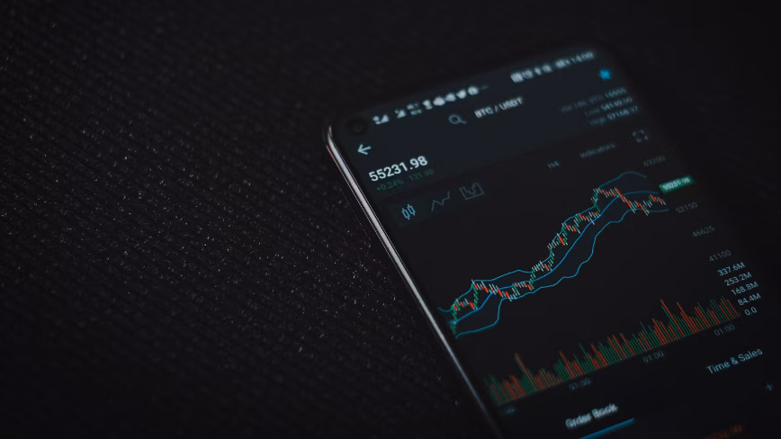 Smartphone on a dark textured surface showing a cryptocurrency candlestick price chart in a trading app.