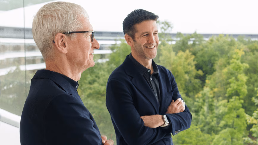 Tim Cook and John Ternus standing together on a balcony at Apple Park, trees behind them, both in dark jackets