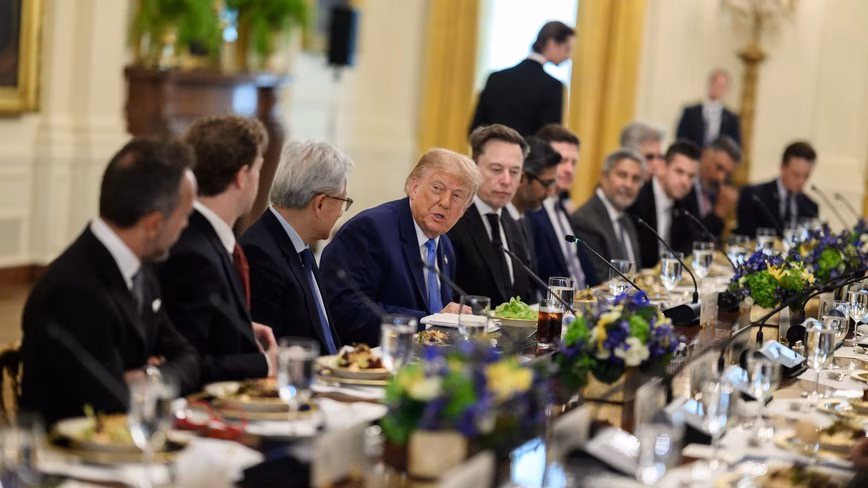 President Donald Trump talking with technology executives seated along a long table with flowers in the East Room of the White House.