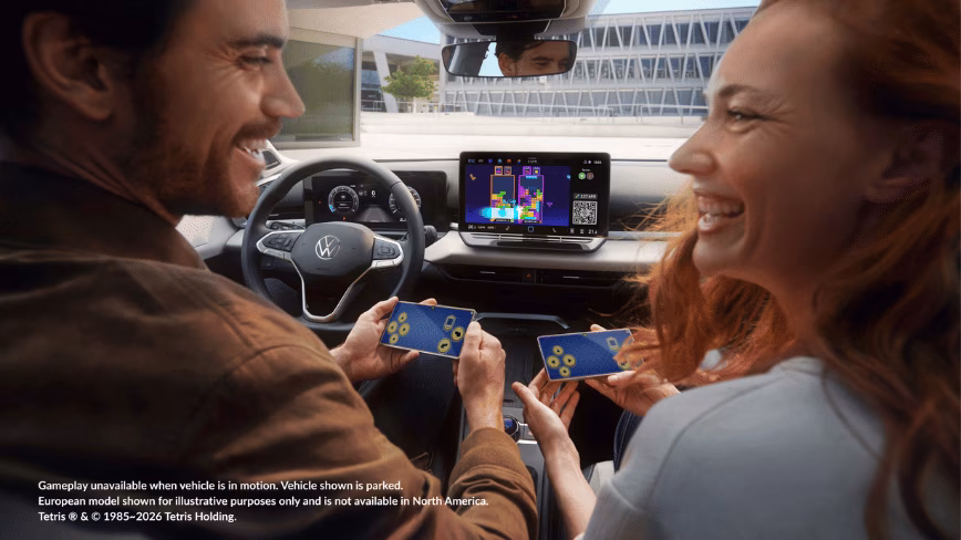 Two people in the front seats of a parked Volkswagen, smiling at each other and holding smartphones as controllers while a game of Tetris runs on the infotainment display