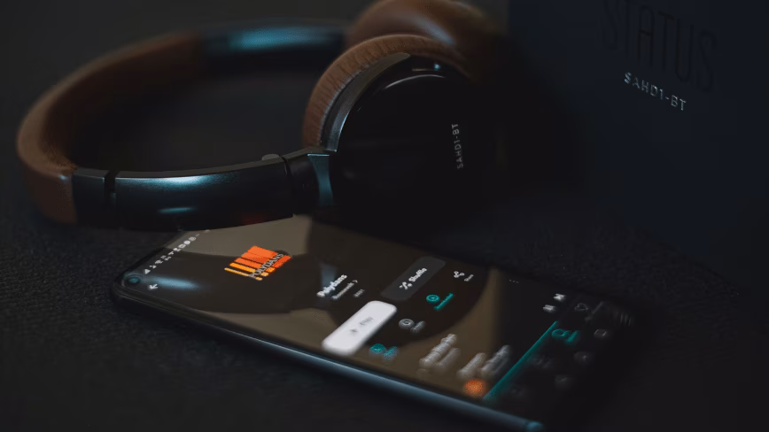 Brown and black over-ear wireless headphones beside a smartphone showing a music streaming app, with the headphones' box on a dark surface