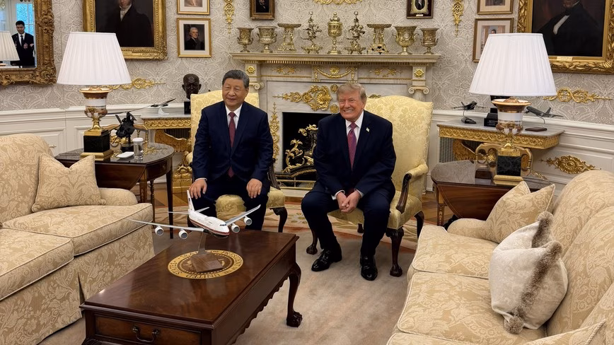 Xi Jinping and Donald Trump seated in gold armchairs before a fireplace in the Oval Office, beside a model of Air Force One