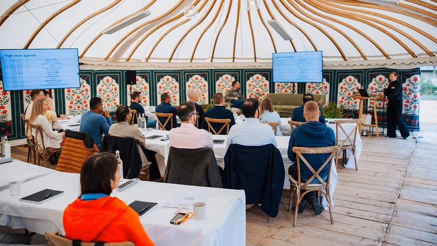 Alex Maxwell of Zapier speaking into a microphone to attendees at tables in a yurt, with Philip Lakin seated on a sofa and two screens showing the day's agenda