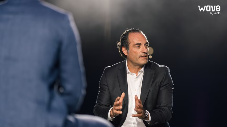 Antonio Filosa, in a dark suit and white shirt with a headset microphone, gestures with both hands as he speaks, a man in a blue jacket blurred in the foreground