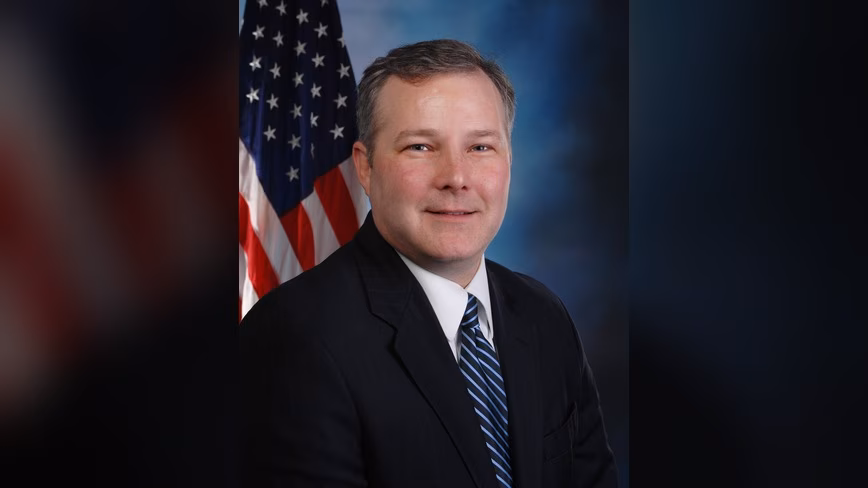 Tim Griffin in a dark suit and striped blue tie, smiling, in front of a US flag and a blue studio backdrop
