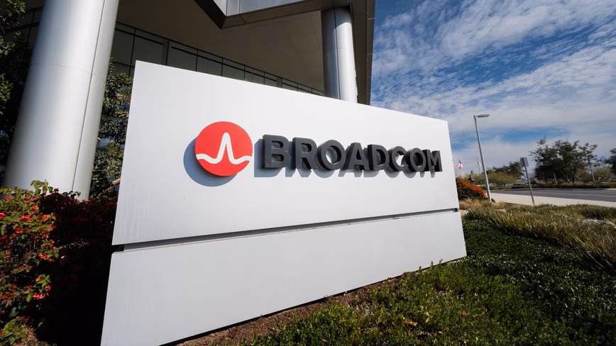 White Broadcom sign with the red logo outside a glass and steel office building, flowering shrubs below and a blue sky with clouds