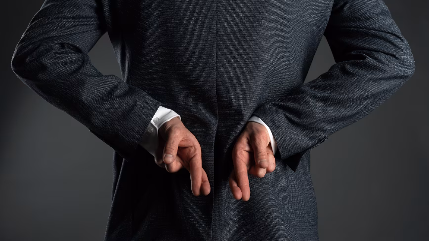 A man in a suit with his fingers crossed behind his back, seen from behind.