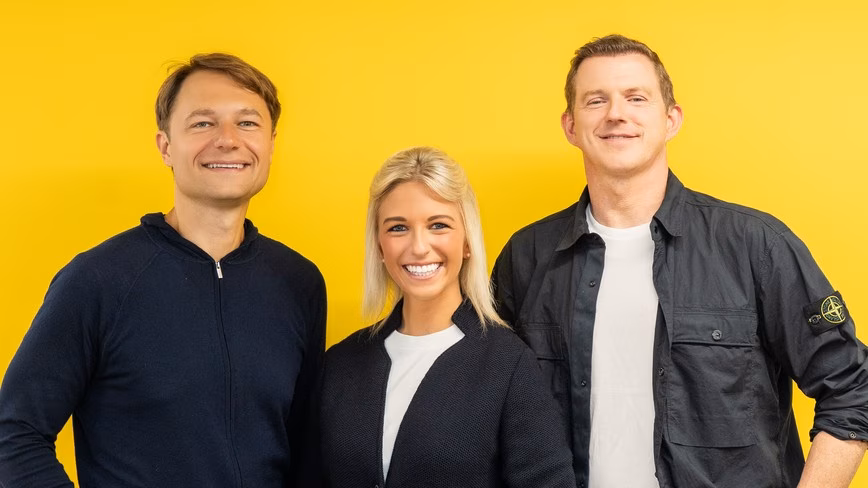 Rytis Vitkauskas, Melissa Klinger and Ross Mason standing together in front of a bright yellow wall