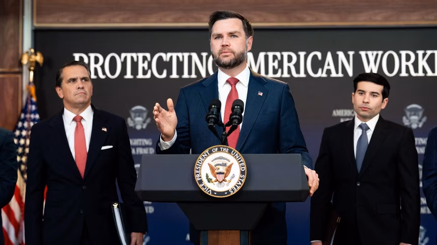 JD Vance gestures while speaking at a podium bearing the vice-presidential seal, with two men in suits standing behind him