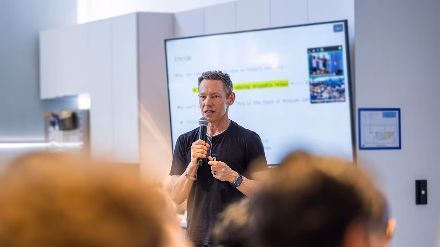 Jeff Holden in a black T-shirt speaking into a microphone in front of a presentation screen, with blurred heads of listeners in the foreground