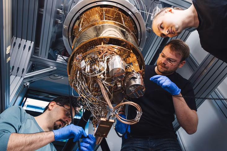 Three researchers in blue gloves work on the gold-plated wiring stages of an open dilution refrigerator