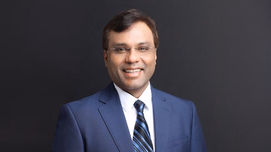 Rohit Prasad smiling in rimless glasses, a navy suit, white shirt and blue striped tie, against a dark grey background