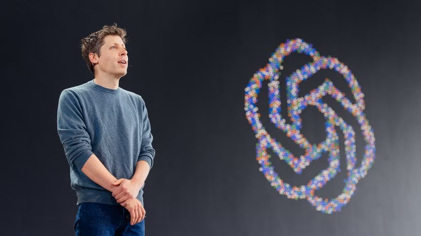 Sam Altman in a grey jumper stands on a dark stage next to a large OpenAI logo made of coloured dots