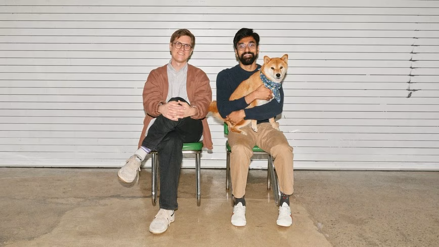 Two smiling men sit on green chairs against a white slatted wall, one holding a Shiba Inu dog wearing a blue bandana