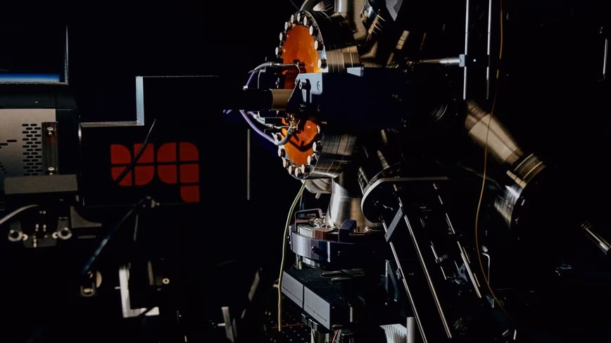 Steel vacuum chamber with an orange viewport and bolted flanges, beside a camera and a box marked with a red UQ logo, in a dark lab