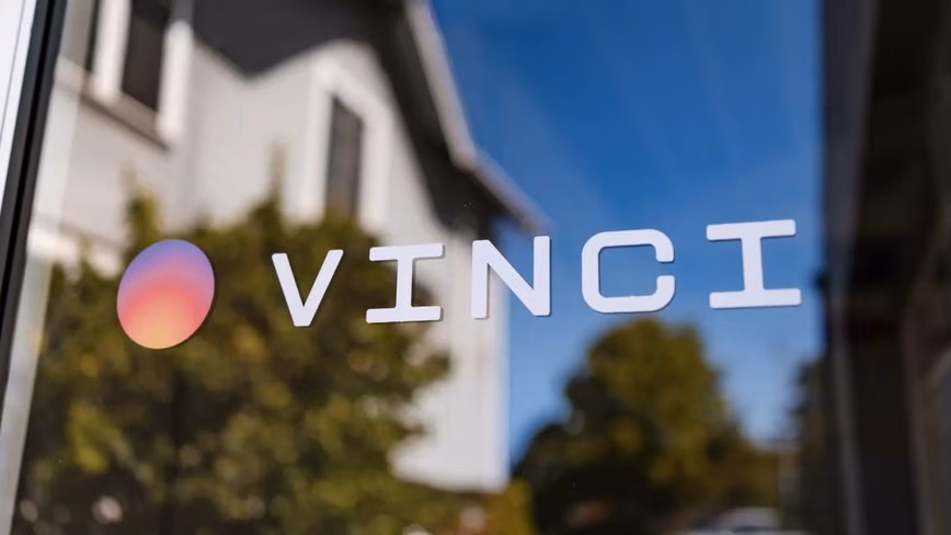 Vinci logo printed on a glass window, reflecting a white building, trees and blue sky.