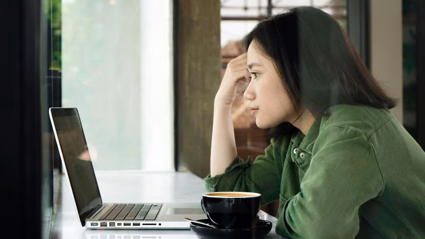 Woman in a green jacket resting her head on her hand as she looks at an open laptop, with a black cup of coffee beside her at a café window counter