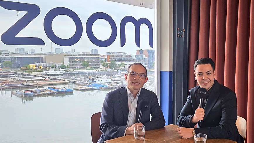 Zoom CTO XD Huang and TNW's Cristian Dina holding Rode microphones at a table, under a Zoom logo on a window over Amsterdam's waterfront